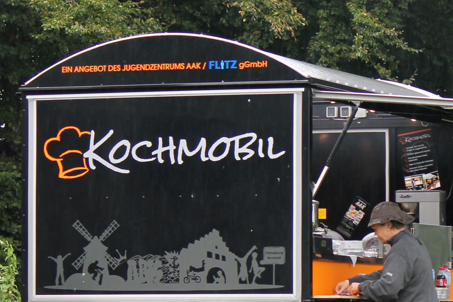 Das Kochmobil ist ein Autoanhänger eines Jugendzentrums. Es sieht wie ein Verkaufsstand auf dem Wochenmarkt mit eingebauter Küche aus und ist mobil einsetzbar.