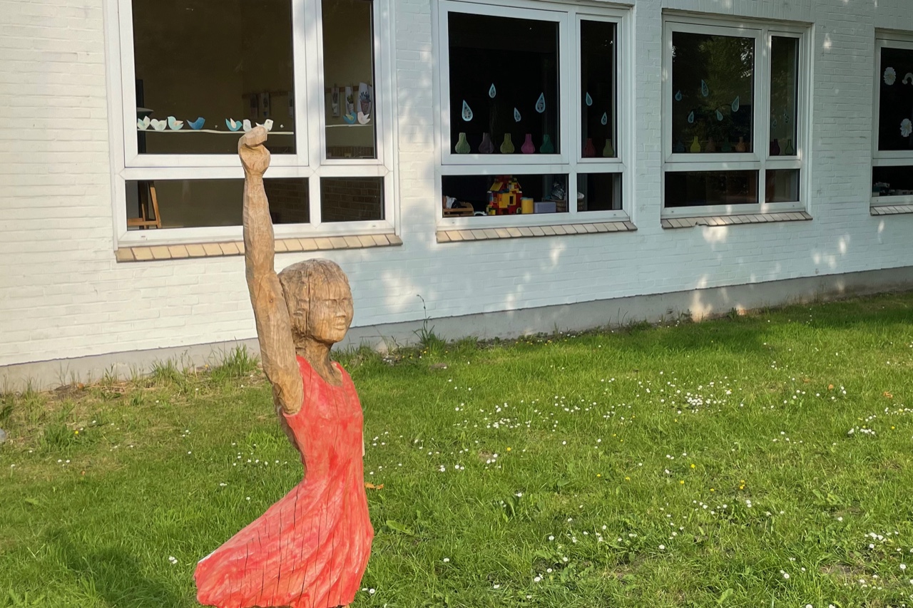 Eine 1,50 Meter große Holzfigur vor den Fenstern einer Grundschule zeigt ein Mädchen im roten Kleid, das sich meldet