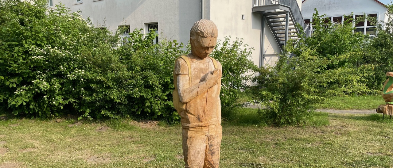 Eine 1,50 Meter große Holzfigur zeigt einen Jungen vor einer Schule, der auf ein Handy schaut