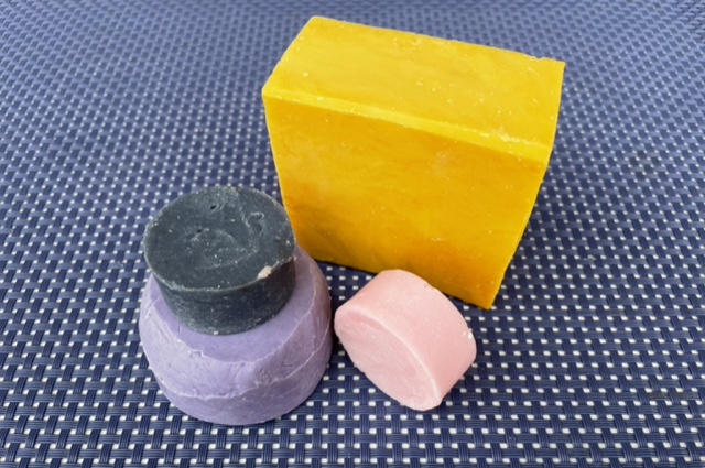 Shampoo-Seifen in Form eines großen, gelben Quaders und drei runde Stücke fliederfarben und rosa, die in kleine Dosen passen
